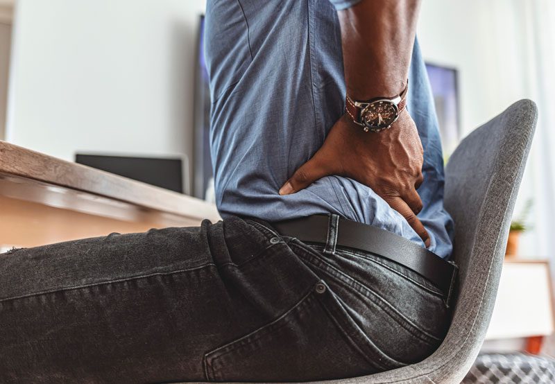 Man-holding-lower-back-while-sitting-in-chair Man-holding-lower-back-while-sitting-in-chair