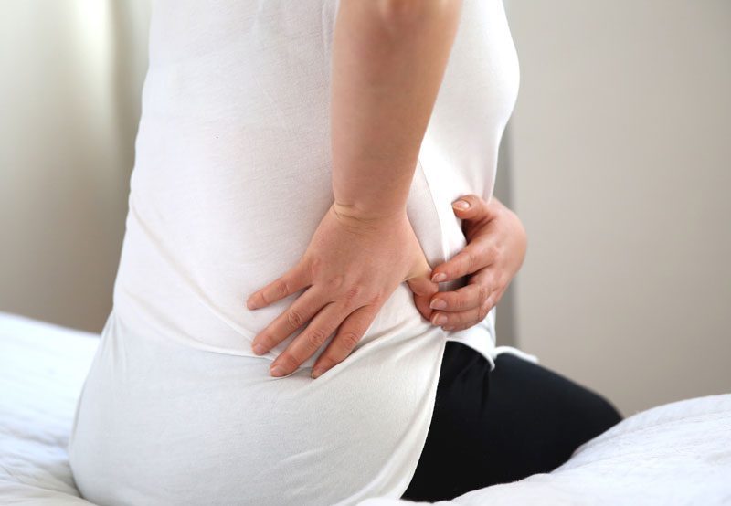 Woman-holding-lower-back-while-sitting-on-bed Woman-holding-lower-back-while-sitting-on-bed